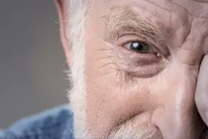 An old man with white hair and a beard is looking at something, his right hand is on his cheek.