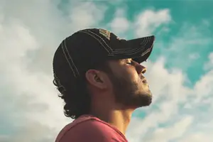A man with long hair and a beard wearing a baseball cap and red shirt looking up at the sky.