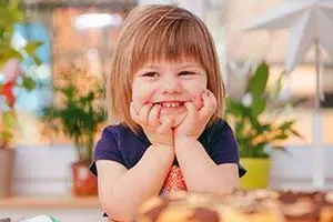 A smiling girl holding her hands on her cheeks in front of a table with plants and a cake.