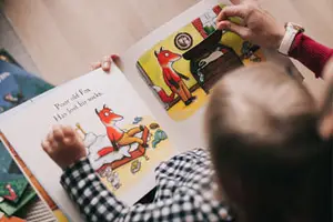A young child is sitting on the floor reading a book with a person holding the book open and pointing to a picture of a fox.
