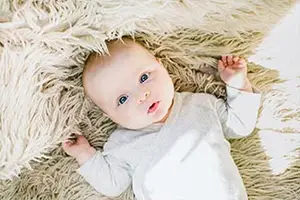 A baby lying on a fluffy white carpet with its hands up and eyes wide open