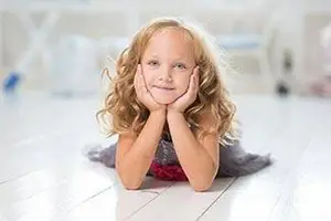 Blonde-haired girl lying on white floor, smiling, and looking at the camera with her hands on her cheeks