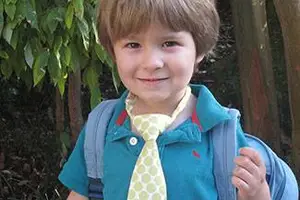 A young boy in a blue shirt and yellow polka-dot tie stands outside, carrying a backpack and smiling for the camera