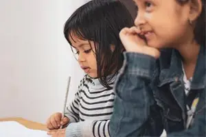 Two young girls sitting at a desk, one writing and the other watching.
