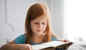 A girl with long red hair is sitting on a bed while reading a book