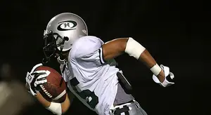 Football player wearing a gray helmet, a white jersey with the number 3 on it, and a black belt with the letter H on it is running while holding a football with both hands. He is wearing a belt and a pair of gloves.