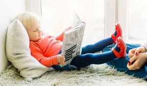 A toddler in red shoes is sitting on a carpet and reading a book with a white pillow beside her and a person sitting on the right side of the toddler.