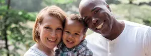 A smiling family of two parents and a child standing in front of a green background