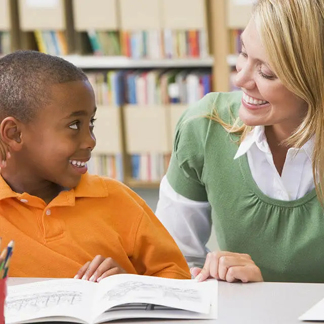 A woman smiles while looking at a boy reading a book in a classroom.