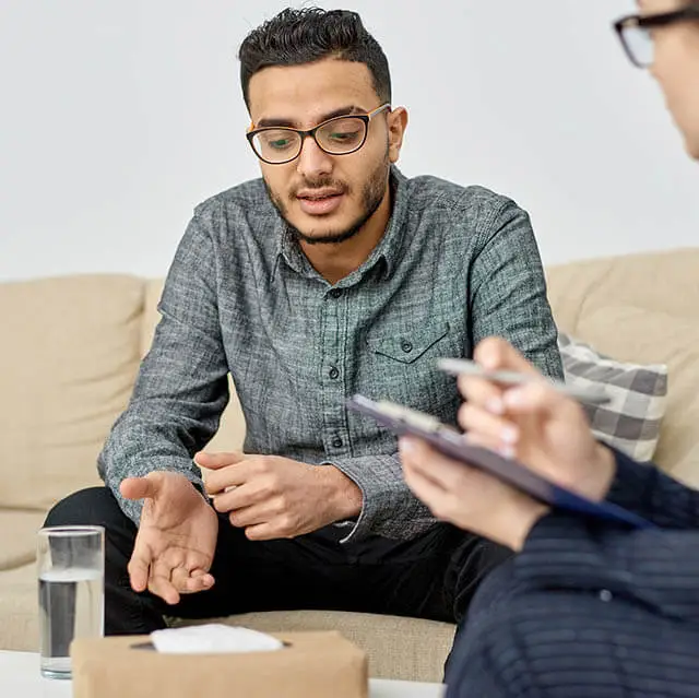 A man is sitting on a couch and talking to a person with a pen and notepad in front of him.