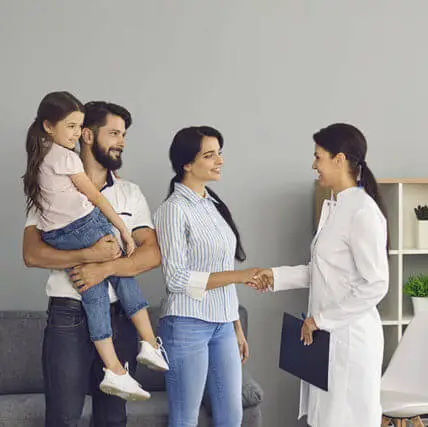A family of four in a living room with a woman in a white lab coat shaking hands with a man holding a girl and a woman holding a folder and a pen.