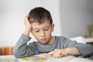 A boy is sitting in a chair with his head on his hand and his other hand pointing to a book on the table.