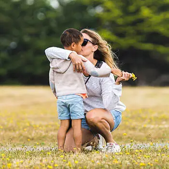 A woman in a white sweatshirt and denim shorts is kneeling in a grassy field with a young boy wearing a light colored hoodie and blue shorts. The woman is holding the child's arm and kissing his cheek.