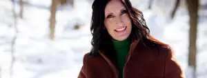 A smiling woman in a green turtleneck and brown jacket stands in a snowy forest with trees behind her.
