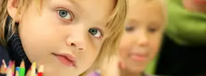 Two children sitting in a classroom one is looking at a row of colored pencils while the other is looking to the side