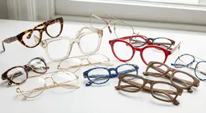 A variety of eyeglasses including tortoise shell, clear, red, and blue frames are displayed on a white surface in front of a window.