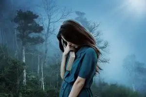 A girl in a blue dress standing in the middle of a foggy forest with her hand covering her face