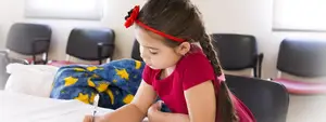 A girl in a red shirt and headband sits at a table in a classroom writing on paper with a pen