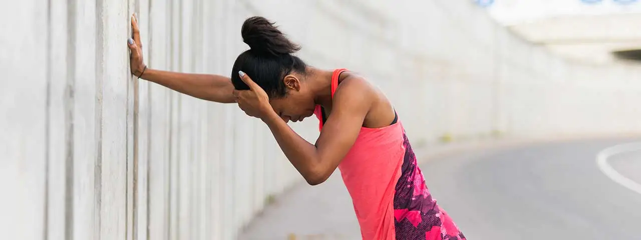 Young woman with black hair wearing pink tank top leaning against wall holding head with one hand and resting hand on wall