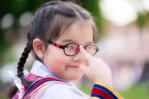 A young girl with glasses, braided hair, and a white shirt with colorful ribbons around her neck is standing on a grassy field and seems to be posing for a photo while touching her nose with her right hand.