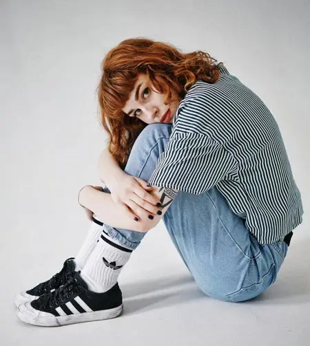 A young woman with red hair is sitting on the floor, wearing a striped shirt, jeans, and sneakers.