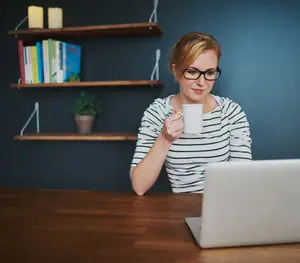 A woman wearing glasses is sitting in front of a desk, smiling and holding a cup of coffee while looking at a laptop on the table in front of her. Behind her is a wooden shelf with books, a plant, and two lit candles. The wall is painted in a dark blue color.