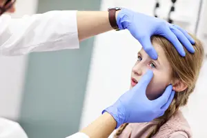 A doctor wearing a watch and blue gloves examines a young girl's ear, likely in a clinic.