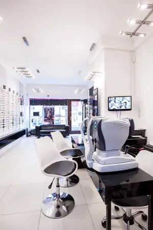 A clean and well-lit optometrist's office with white chairs, a black desk, and an eye examination machine on top, along with ceiling lights and a television on the wall.