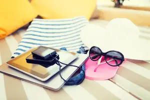 Electronic devices like a tablet and a cell phone are kept on a striped cloth along with some other objects like a hat, a pair of glasses, and a pair of slippers.