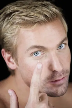 A man with short blonde hair and blue eyes is smiling and pointing at his eye while wearing a black top.
