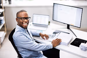 A smiling man sitting at a desk with a laptop and calculator in an office.