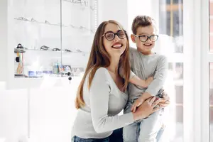 A woman and a boy wearing glasses are standing together in front of a shelf with eyeglasses inside a store
