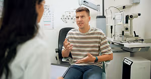 A man wearing a striped shirt is sitting on a chair and talking to a woman who is looking at a monitor. Behind them is a black chair, a machine, and a wall with an eye chart.