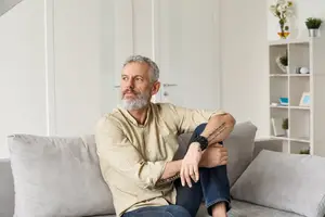An adult male is sitting on a couch with his arms crossed and looking at something in the room.
