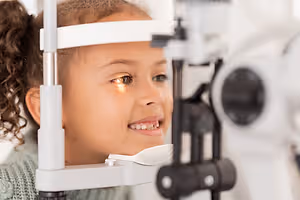 A young girl wearing glasses is looking through an eye examination device with a smiling face.