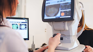 A person adjusting a machine with a monitor screen in front of them, while another person is sitting behind them.