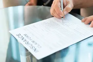 A person fills out an insurance claim form with a pen on a glass table.