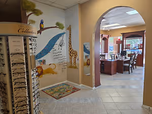 A room with a table, chairs, computer, shelves, and a carpet on the floor, with a wall featuring a giraffe, lion, and other animals, and a display of eyeglasses labeled "The Exclusive Collection".