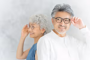 Two elderly people, a man and a woman, are standing together in front of a white wall, smiling and wearing glasses.