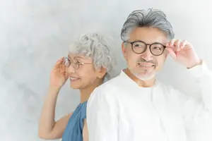 Two elderly people, a man and a woman, are standing together in front of a white wall, smiling and wearing glasses.
