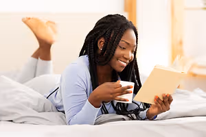 A woman is lying on a bed with her feet up, smiling and holding a book and a cup.