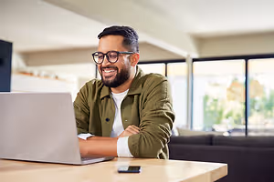 A man in a green jacket with glasses and a beard is sitting at a desk with a laptop and a cell phone.