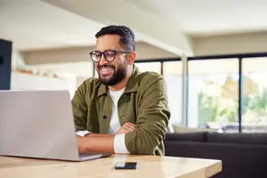 A man in a green jacket with glasses and a beard is sitting at a desk with a laptop and a cell phone.