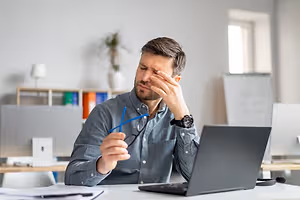 A man sitting at a desk in an office room wearing a blue shirt and holding his head with his left hand.