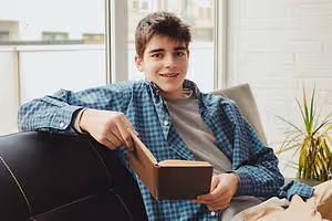 A boy is sitting on a couch and holding a book in his hand, smiling and posing for a photo.