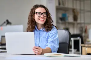 A smiling woman with glasses is sitting at a desk in an office, holding a laptop.