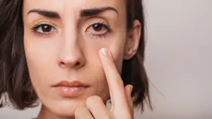 A woman with brown hair is holding her nose with her finger and looking at something in front of her.