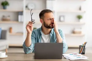 A man is sitting at a desk with his hands on his face. He is wearing a blue long-sleeve shirt and a white undershirt. He is holding a pair of glasses in his right hand. On the desk are a laptop, a cup, a pen holder, a stack of papers, and a potted plant. Behind him are shelves with books, a clock, and a plant.