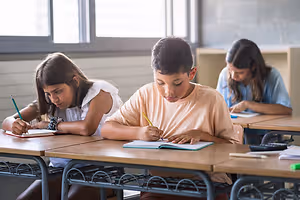 Three students are sitting at desks in a classroom, working on their notebooks with pens, one girl with a bracelet and long hair, a boy with short hair, and a girl with glasses, with sunlight coming through a window in the background.