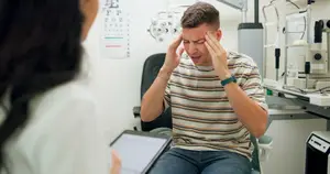 A man sitting in a chair at an optometrist's office with his hands on his head looking at a woman holding a tablet.